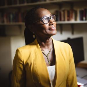 Simone A James Alexander stands in her office wearing a yellow blazer. She smiles and looks away from the camera towards the light streaming in through the window.