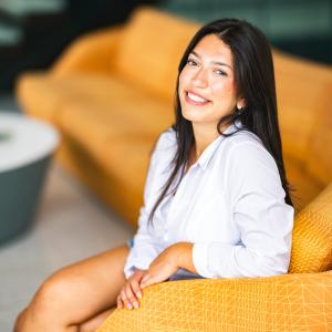 Jordan Kotch sits on an orange sofa and smiles at the camera wearing a white button up shirt.