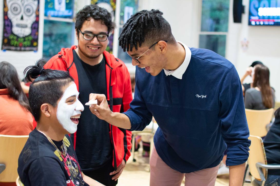 A student paints another student with white face paint to create a skull for a day of the dead celebration 