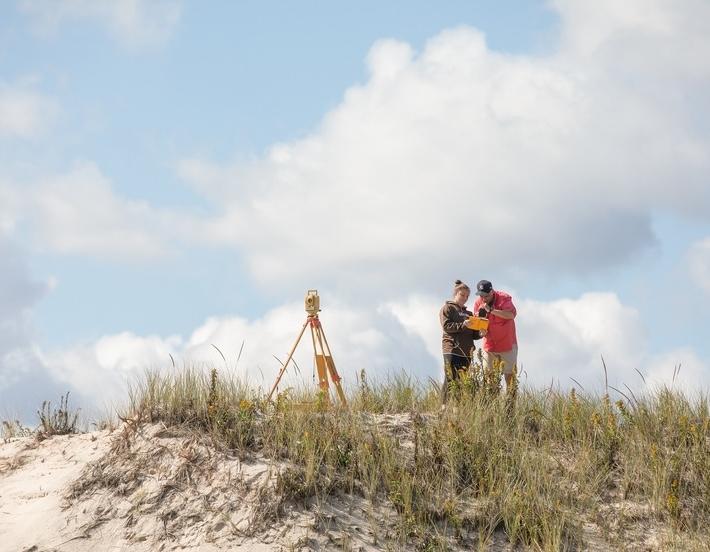 Professor and student stand on a beach dune taking notes