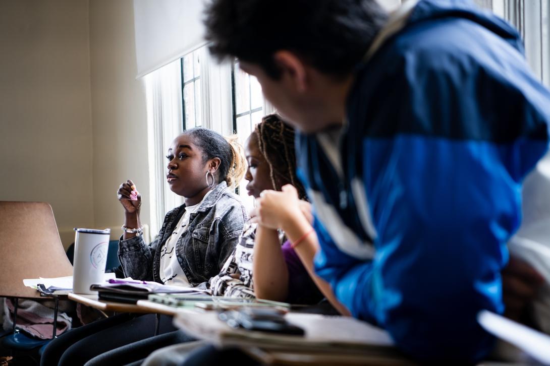 Students sit in a circle discussing with the camera focused on a young Black woman