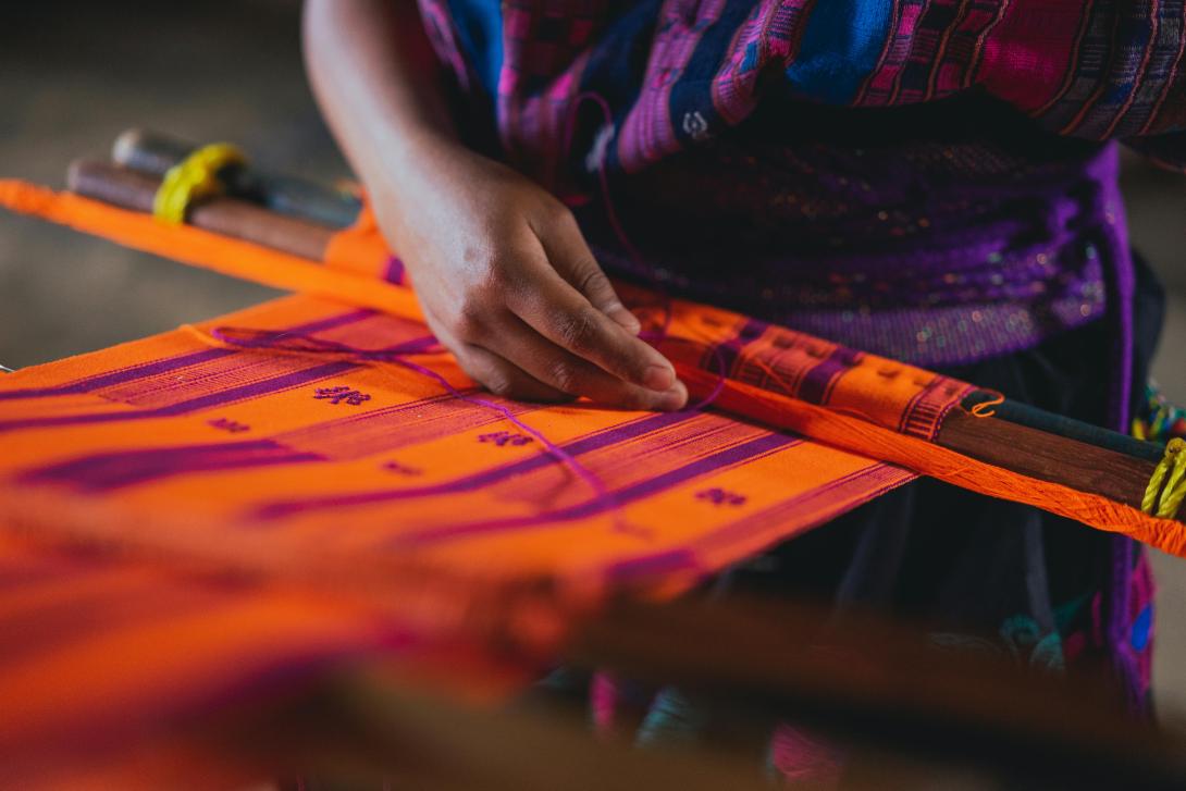 A woman weaves an orange and maroon tapestry 