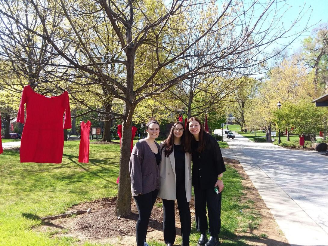 Three students stand next to red dresses hanging from trees