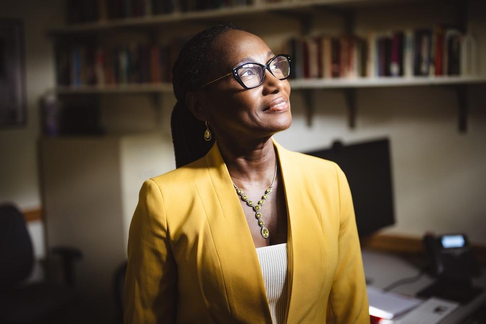 Simone A James Alexander stands in her office wearing a yellow blazer. She smiles and looks away from the camera towards the light streaming in through the window.